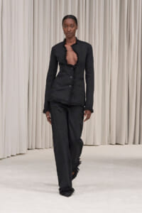 Model walking a fashion runway in a black frayed-edge blazer and matching trousers with a deep open neckline.