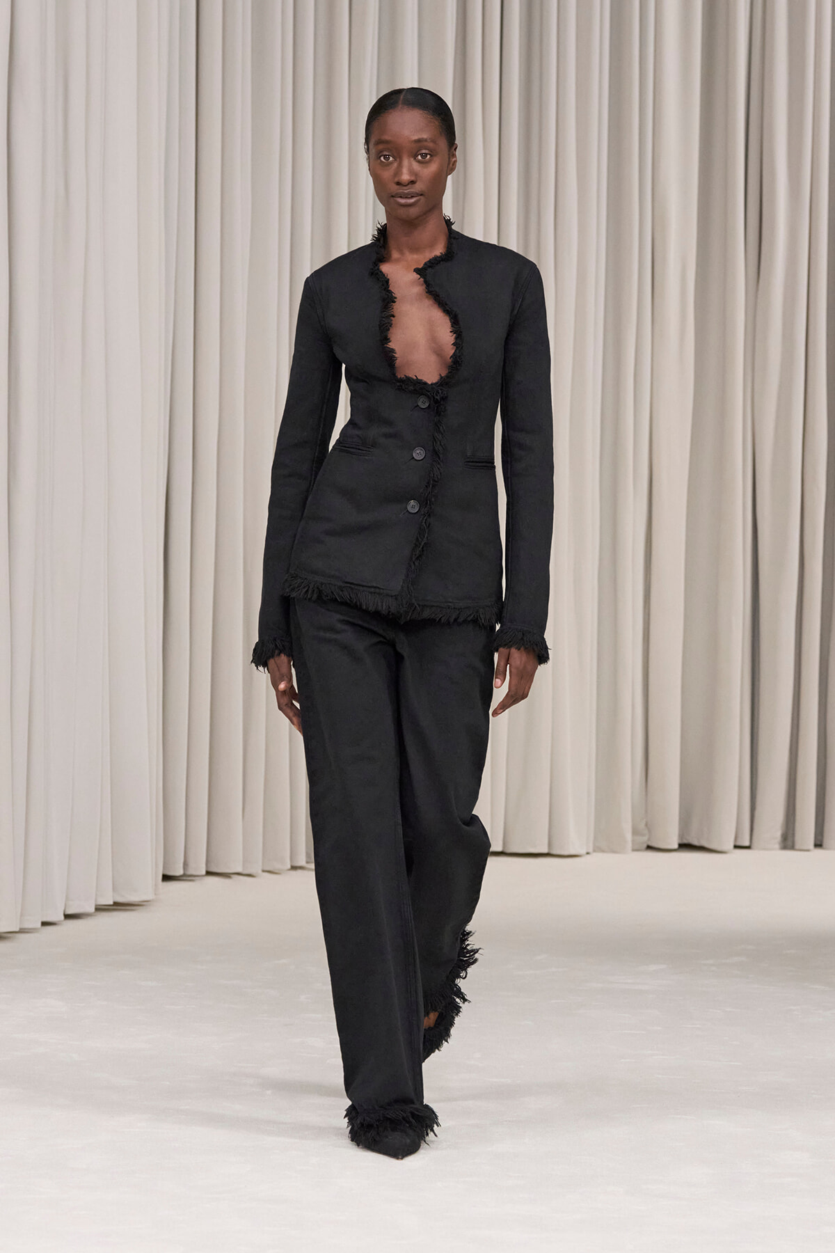 Model walking a fashion runway in a black frayed-edge blazer and matching trousers with a deep open neckline.