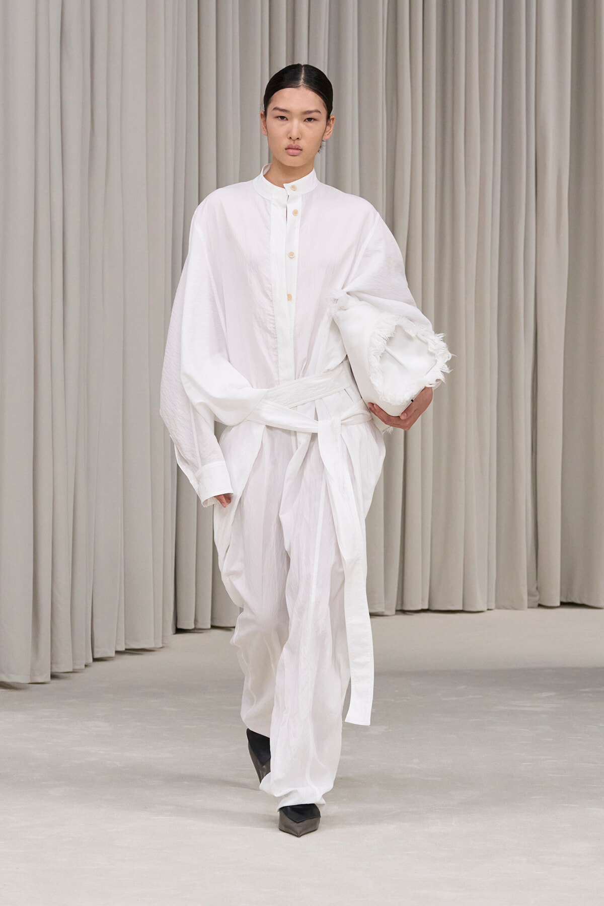 Model on a runway wearing an oversized white shirt and wide-leg white pants, carrying a white clutch against beige curtains.
