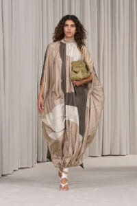 Model wearing a loose, geometric-patterned kaftan in nude tones, holding a beige pouch and wearing white wrap sandals.