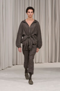 Male model walks the runway in a gray crinkled jumpsuit with a front zipper and large knot at the waist, hooded and tied, neutral backdrop of curtains.