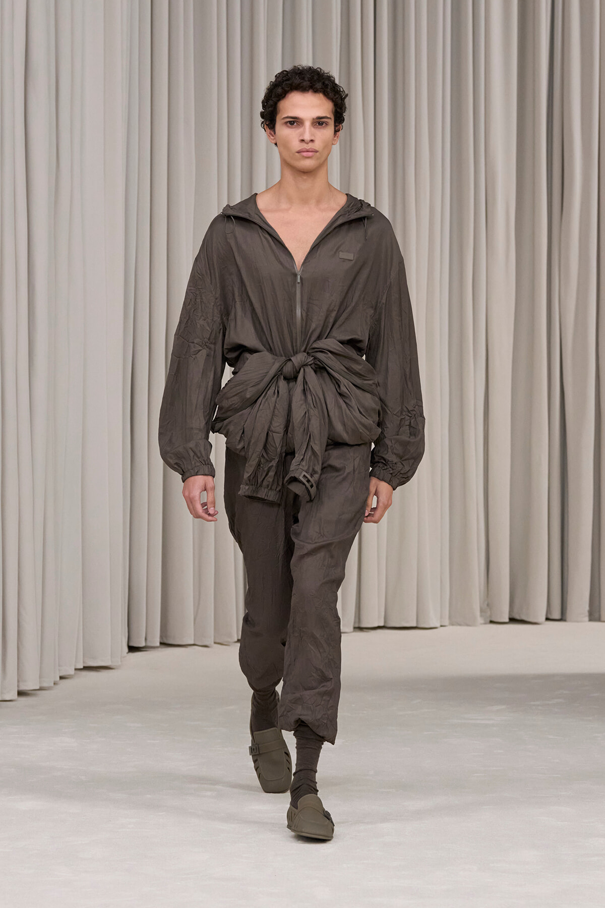 Male model walks the runway in a gray crinkled jumpsuit with a front zipper and large knot at the waist, hooded and tied, neutral backdrop of curtains.