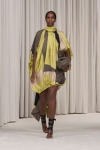 Runway model wearing a loose color-block yellow, beige, and gray dress, carrying a gray handbag and strappy sandals on a white runway backdrop.