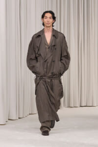Male model walks the runway in an oversized brown trench coat with a waist belt, neutral curtain backdrop behind him.
