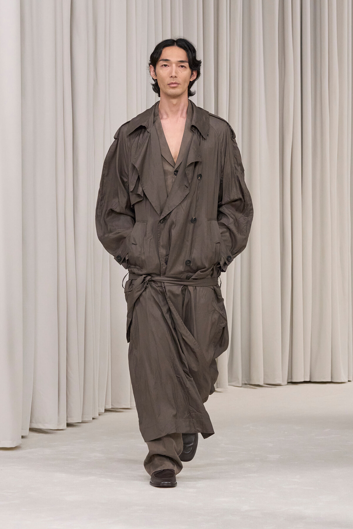 Male model walks the runway in an oversized brown trench coat with a waist belt, neutral curtain backdrop behind him.