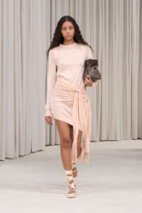 Model walking a runway in a blush pink wrap-dress with a waist knot, carrying a gray handbag.