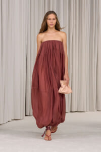 Model walking runway in a strapless, rust-colored, billowy maxi dress, carrying a pale pink clutch and wearing sandals.