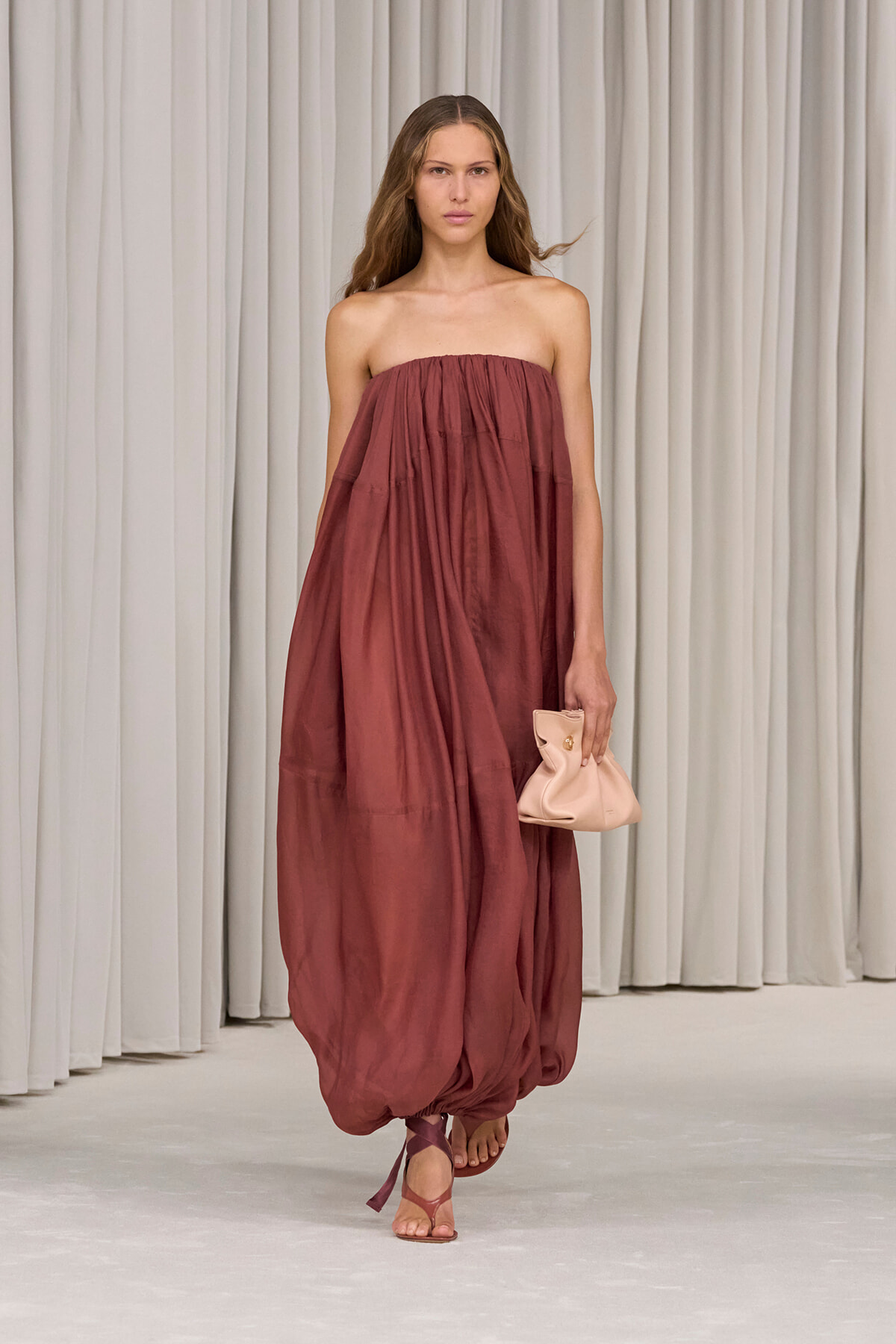 Model walking runway in a strapless, rust-colored, billowy maxi dress, carrying a pale pink clutch and wearing sandals.