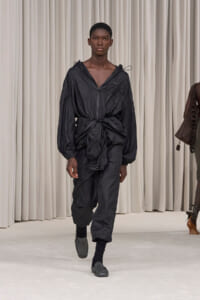 Male model walks the runway in an all-black, oversized, crinkled jumpsuit with a waist-tied knot, black socks and sandals.