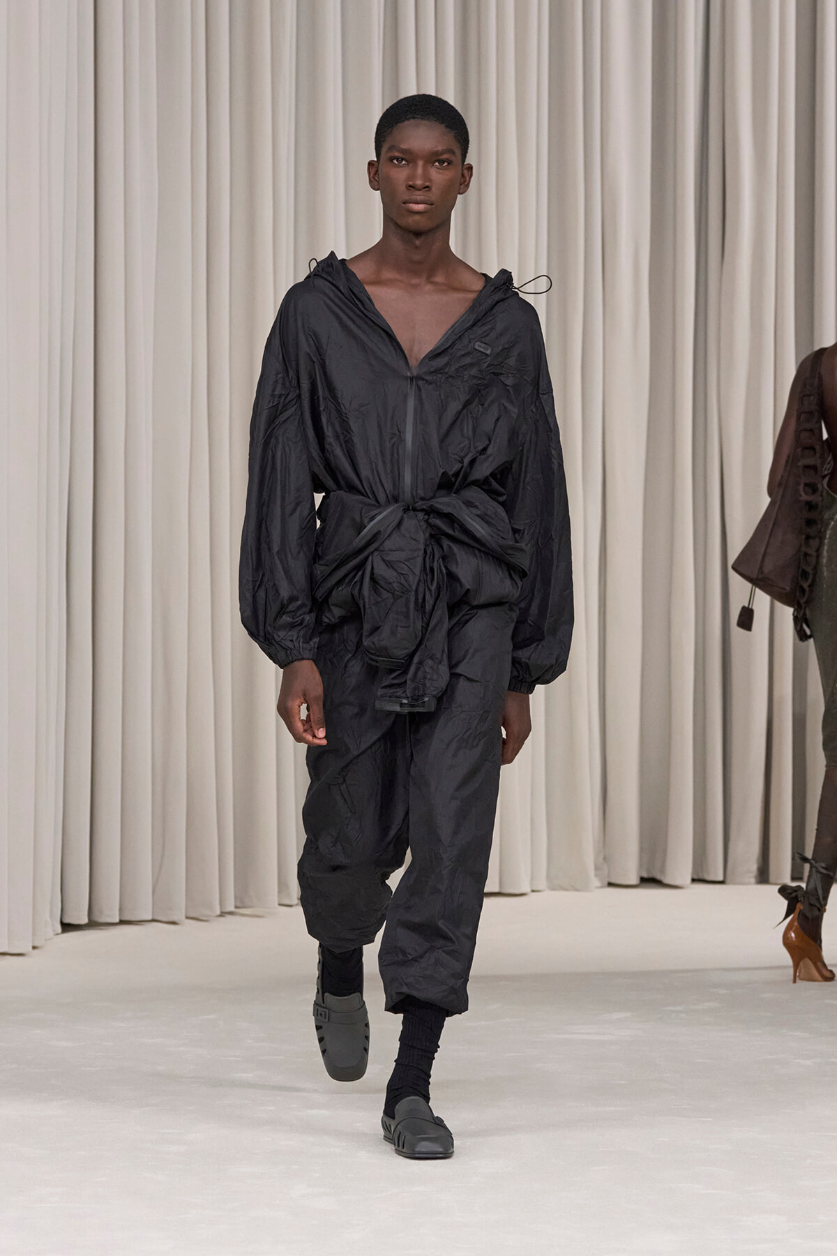 Male model walks the runway in an all-black, oversized, crinkled jumpsuit with a waist-tied knot, black socks and sandals.