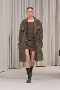 Male model on a runway wearing an olive oversized trench over a brown knit dress, with brown socks and shoes, neutral curtain backdrop at the show.