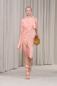 Runway model in a pale pink one-shoulder dress with a waist knot and high slit, holding a tan suede clutch.