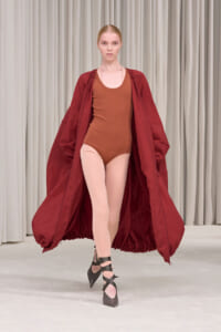 Model on a runway wearing a rust-brown bodysuit under a voluminous burgundy coat, pale tights, and black crisscross heels.