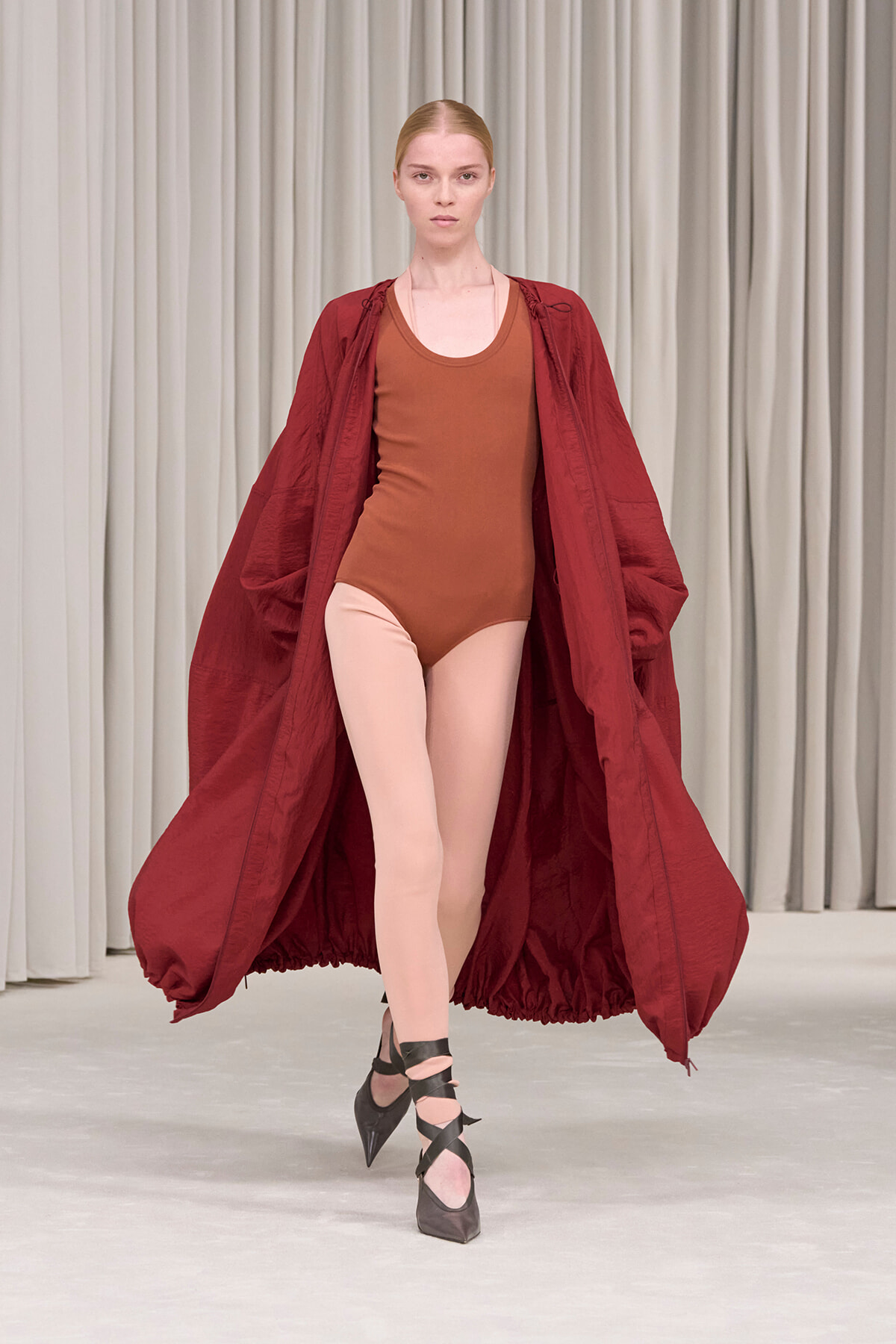 Model on a runway wearing a rust-brown bodysuit under a voluminous burgundy coat, pale tights, and black crisscross heels.