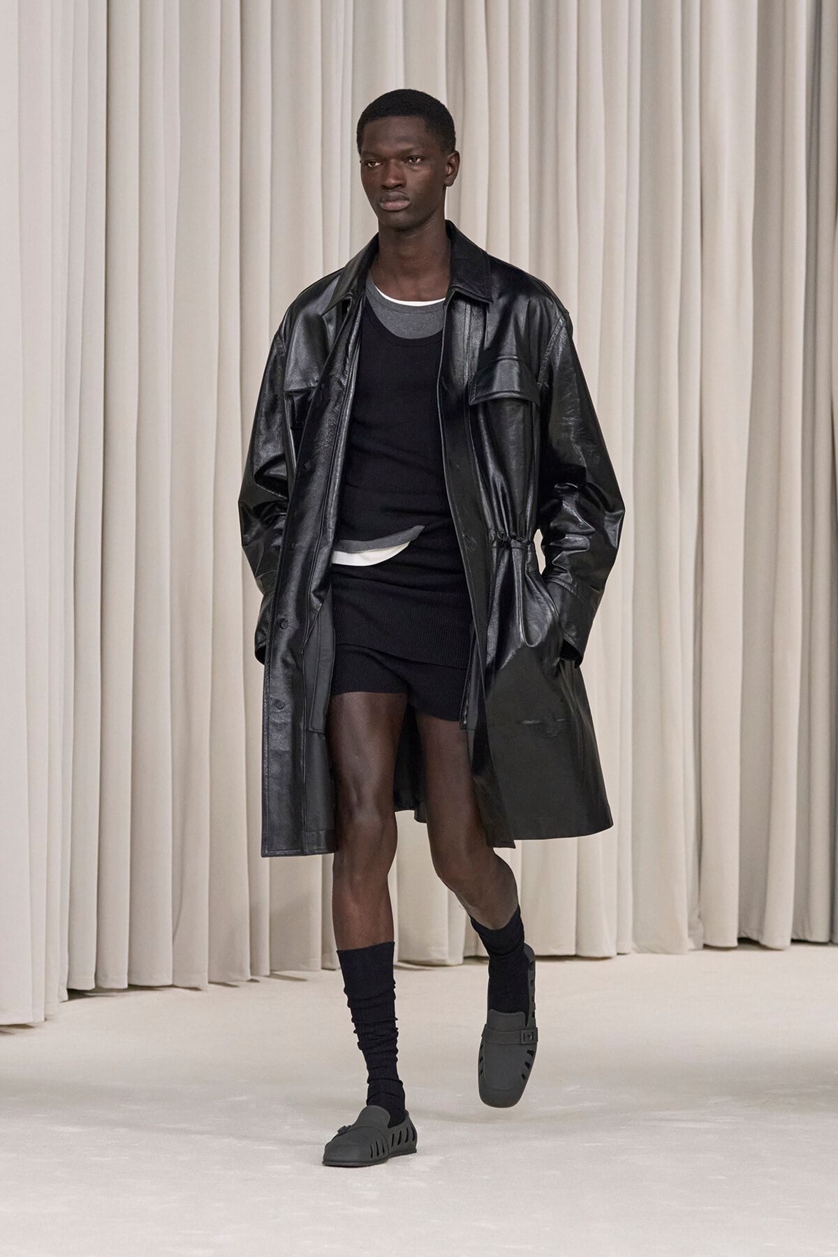 Male model on runway wearing a black oversized leather trench over a black top and shorts, with gray sandals and black socks.