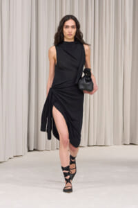 Model walking a runway in a black sleeveless draped dress with a high neckline and a side knot, revealing a leg through a high slit, paired with black strappy heels and a small black handbag.