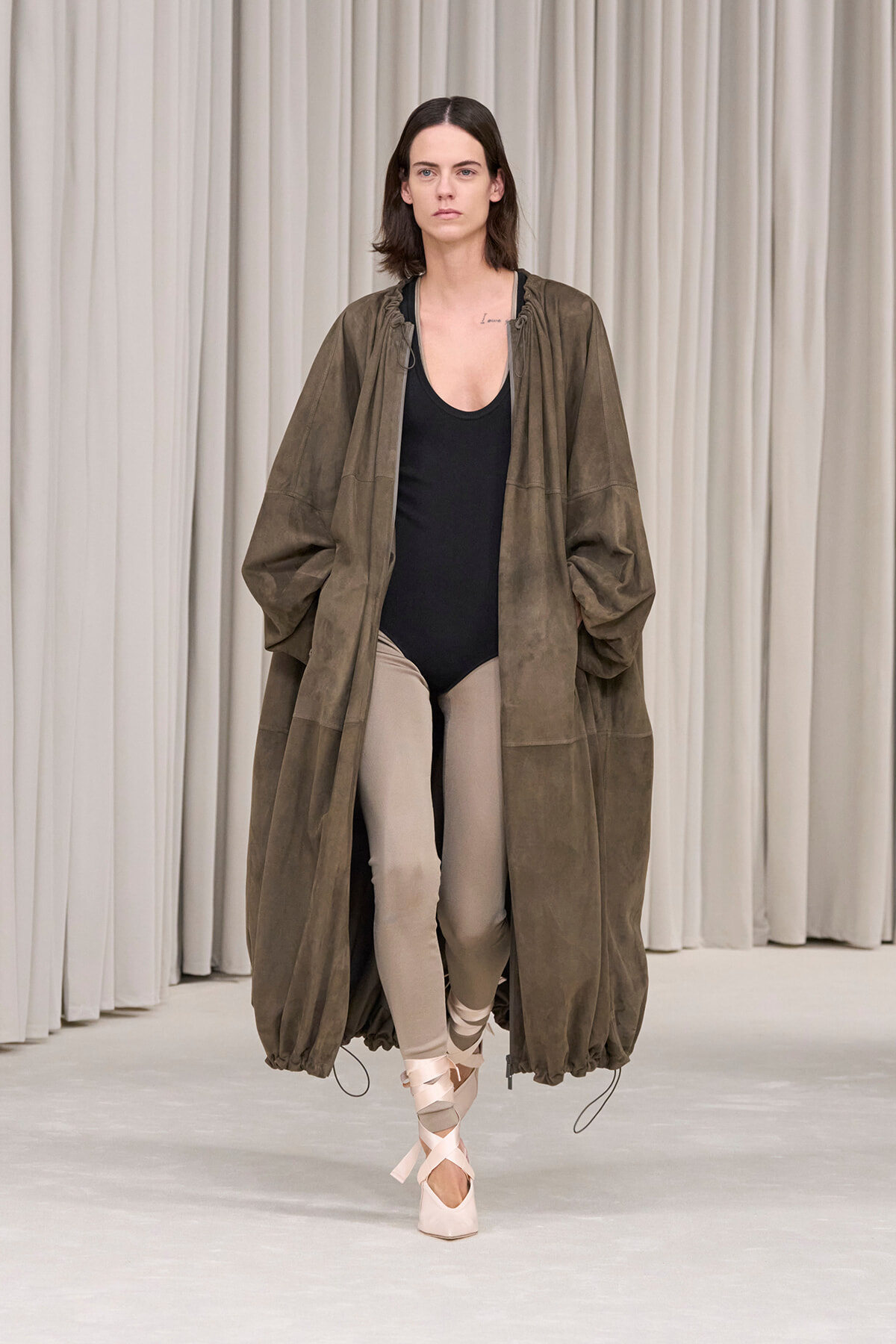 Model walking a runway in an oversized olive-brown coat over a black bodysuit with beige leggings and pink wrap ballet shoes.