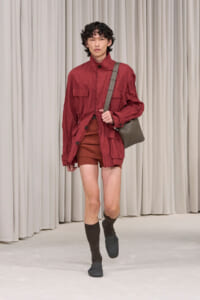 Male model walking a runway in a red utility jacket, brown shorts, dark socks, and sandals, carrying a gray shoulder bag.
