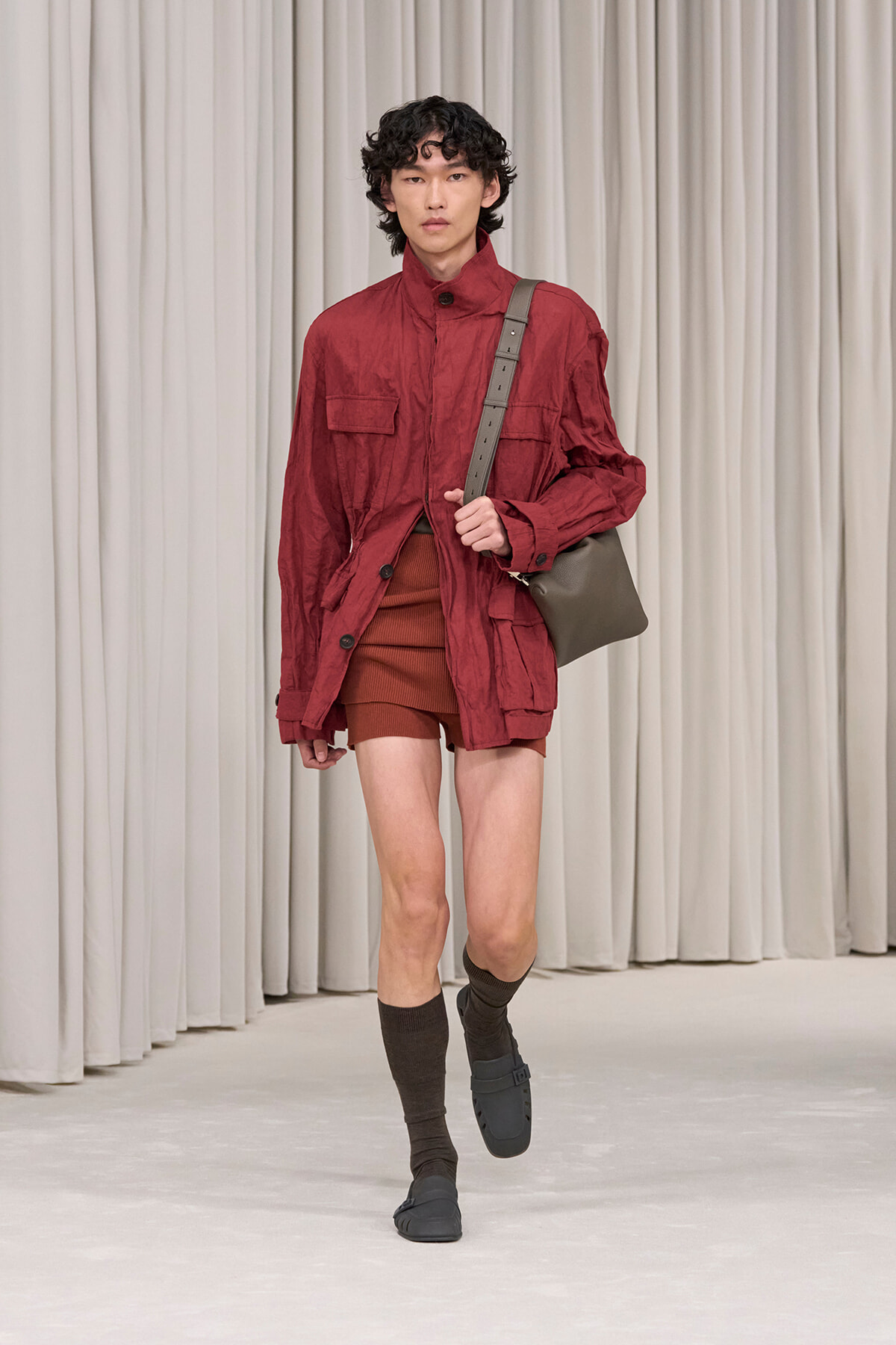 Male model walking a runway in a red utility jacket, brown shorts, dark socks, and sandals, carrying a gray shoulder bag.