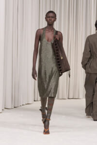 Model walking the runway in a sparkly silver-green halter dress, carrying a large brown strap-laced bag. Boots feature wrapped straps.
