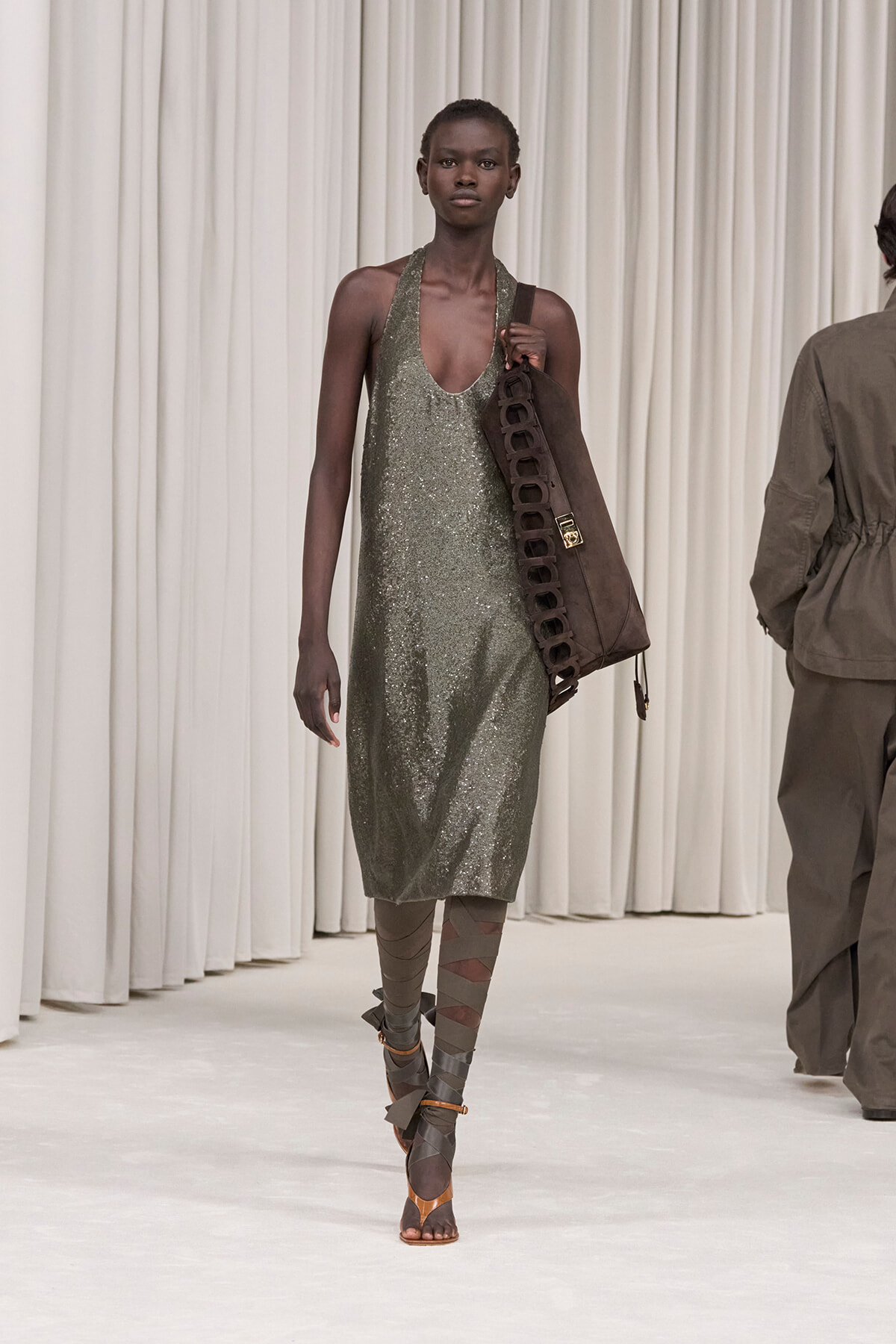 Model walking the runway in a sparkly silver-green halter dress, carrying a large brown strap-laced bag. Boots feature wrapped straps.