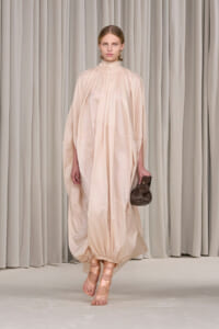 Model on runway in a loose, pale pink-beige floor-length dress with a high neckline and voluminous sleeves, holding a brown clutch.