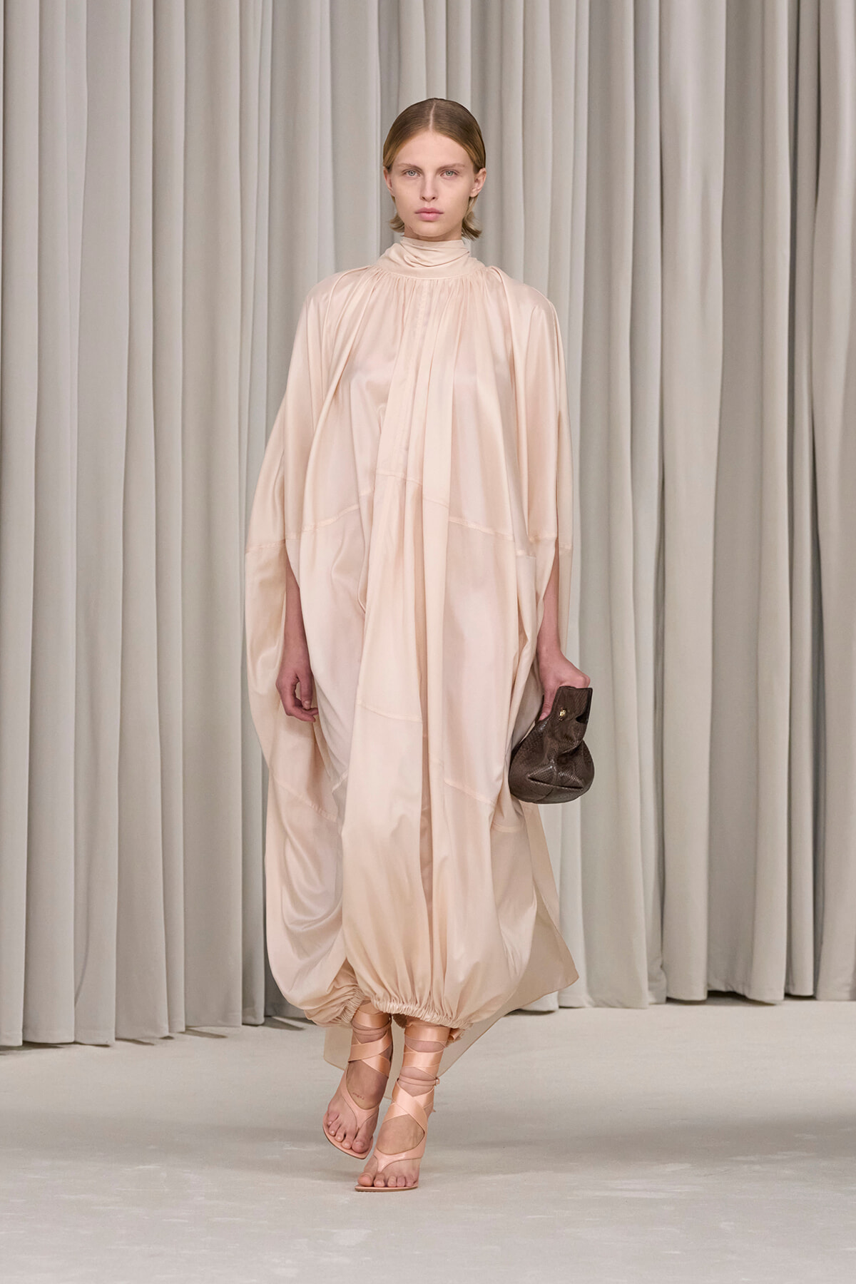 Model on runway in a loose, pale pink-beige floor-length dress with a high neckline and voluminous sleeves, holding a brown clutch.