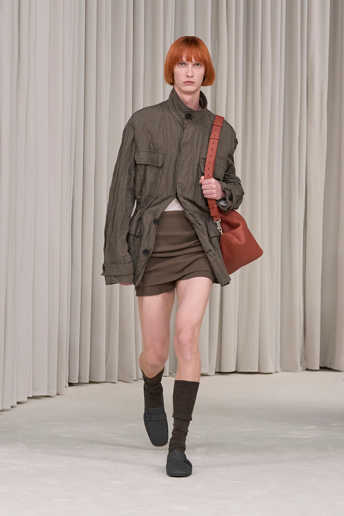 Female model on a runway wearing an oversized olive-green utility jacket, brown ribbed mini skirt, dark socks, and black sandals, with a red shoulder bag.