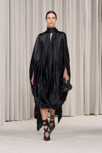 Model walking a runway in a glossy black voluminous satin dress with a high neck, holding a small black handbag and wearing strappy heels.