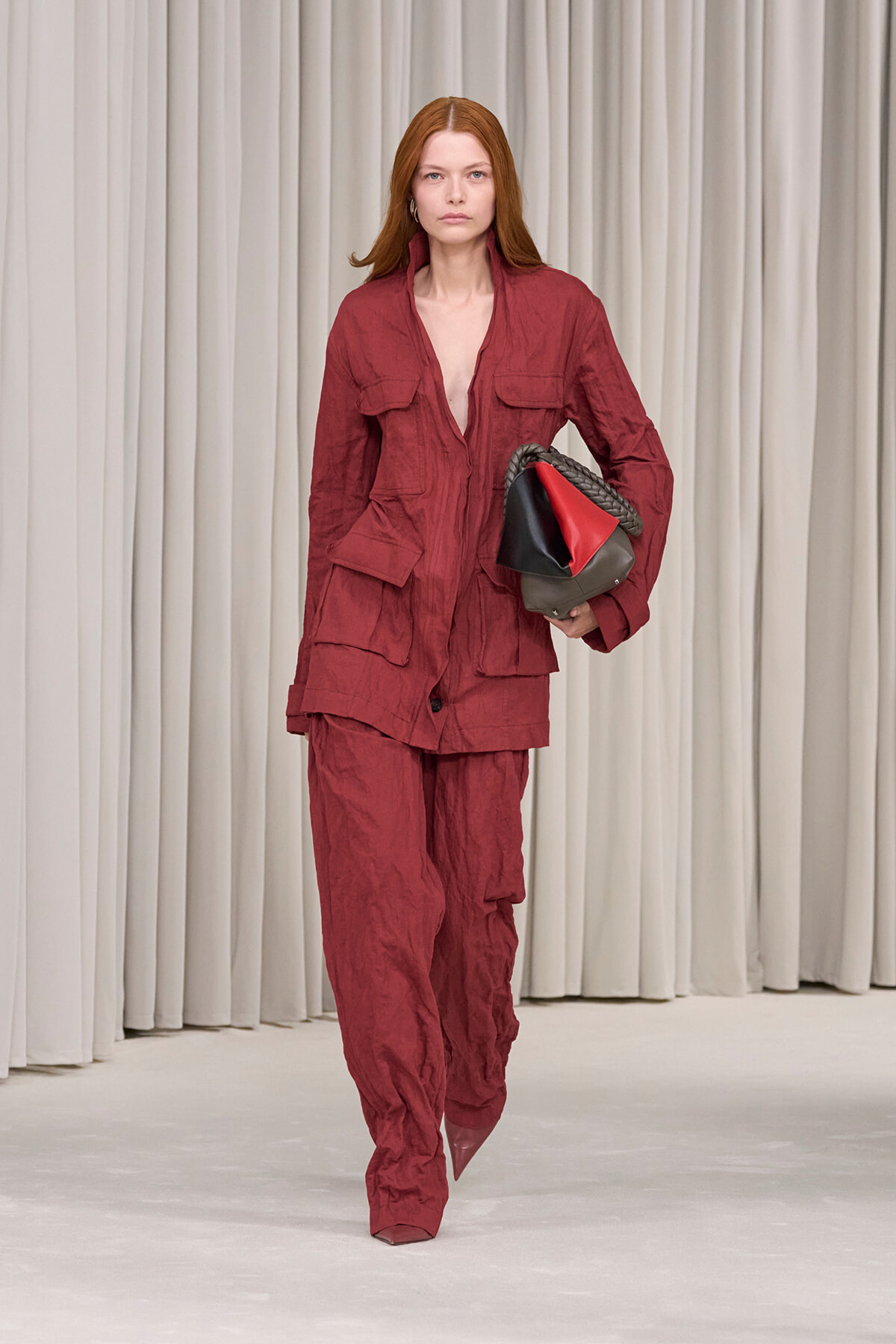 Model walking a fashion runway in a burgundy utility suit with large pockets, carrying a color-block red/black/brown handbag against a pale curtain backdrop.
