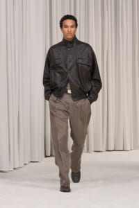 Male model walking the runway in a black leather jacket and taupe-brown pants on a neutral backdrop.