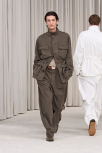 Male model walks a runway in an olive utility jacket with large pockets and matching pants, beige belt, neutral backdrop.