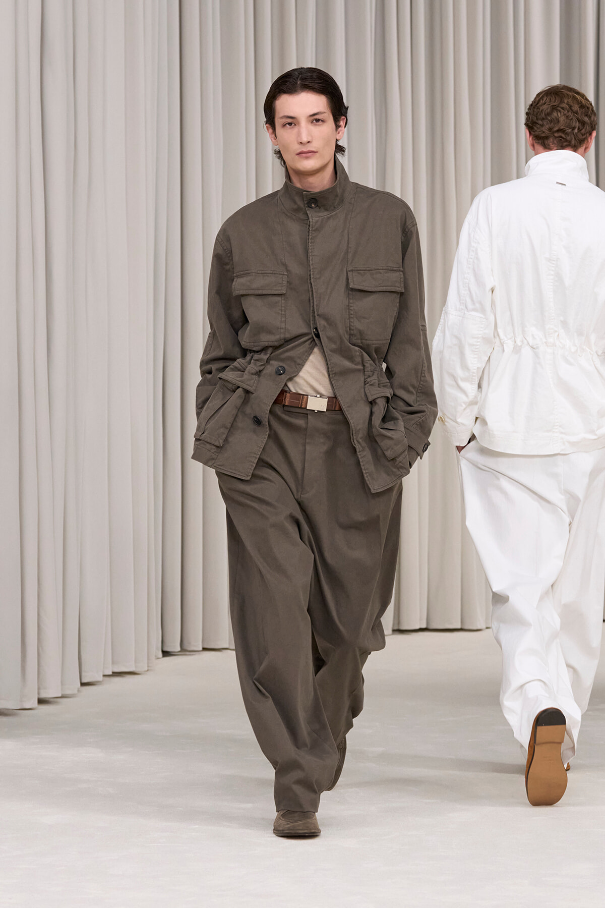 Male model walks a runway in an olive utility jacket with large pockets and matching pants, beige belt, neutral backdrop.