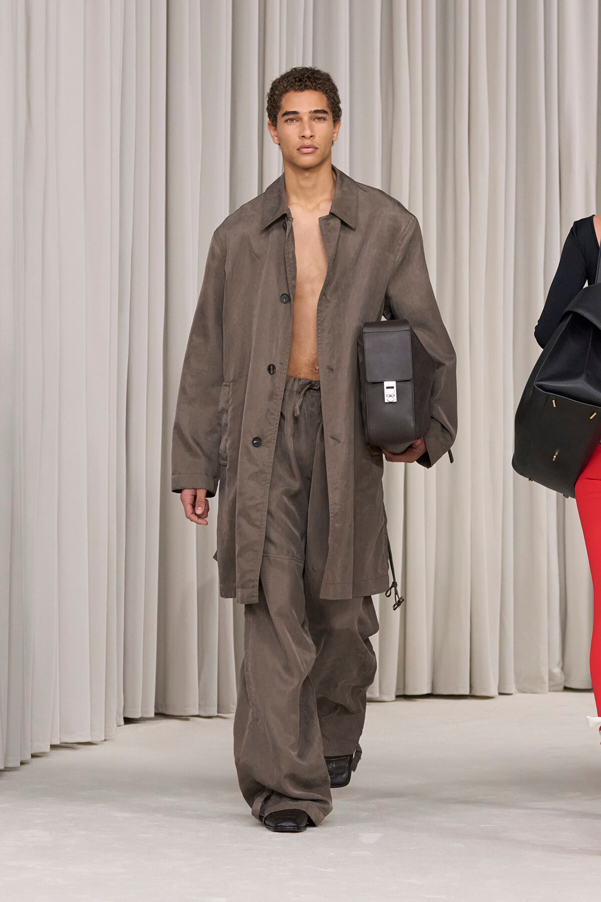 Male model on a runway wearing an oversized taupe coat and matching baggy pants, carrying a black rectangular bag under his arm.