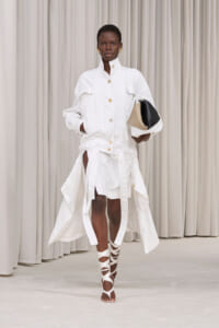 Model in an all-white high-fashion outfit: a button-front, high-collar jacket with gold buttons and a long belted skirt, holding a color-block clutch and wearing white wrap sandals against a neutral backdrop.