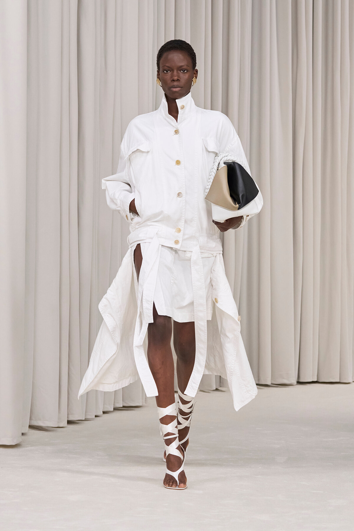 Model in an all-white high-fashion outfit: a button-front, high-collar jacket with gold buttons and a long belted skirt, holding a color-block clutch and wearing white wrap sandals against a neutral backdrop.