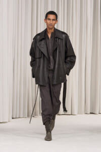 Male model walking a fashion runway in an oversized black leather trench and dark layered outfit, hands in pockets, neutral expression.