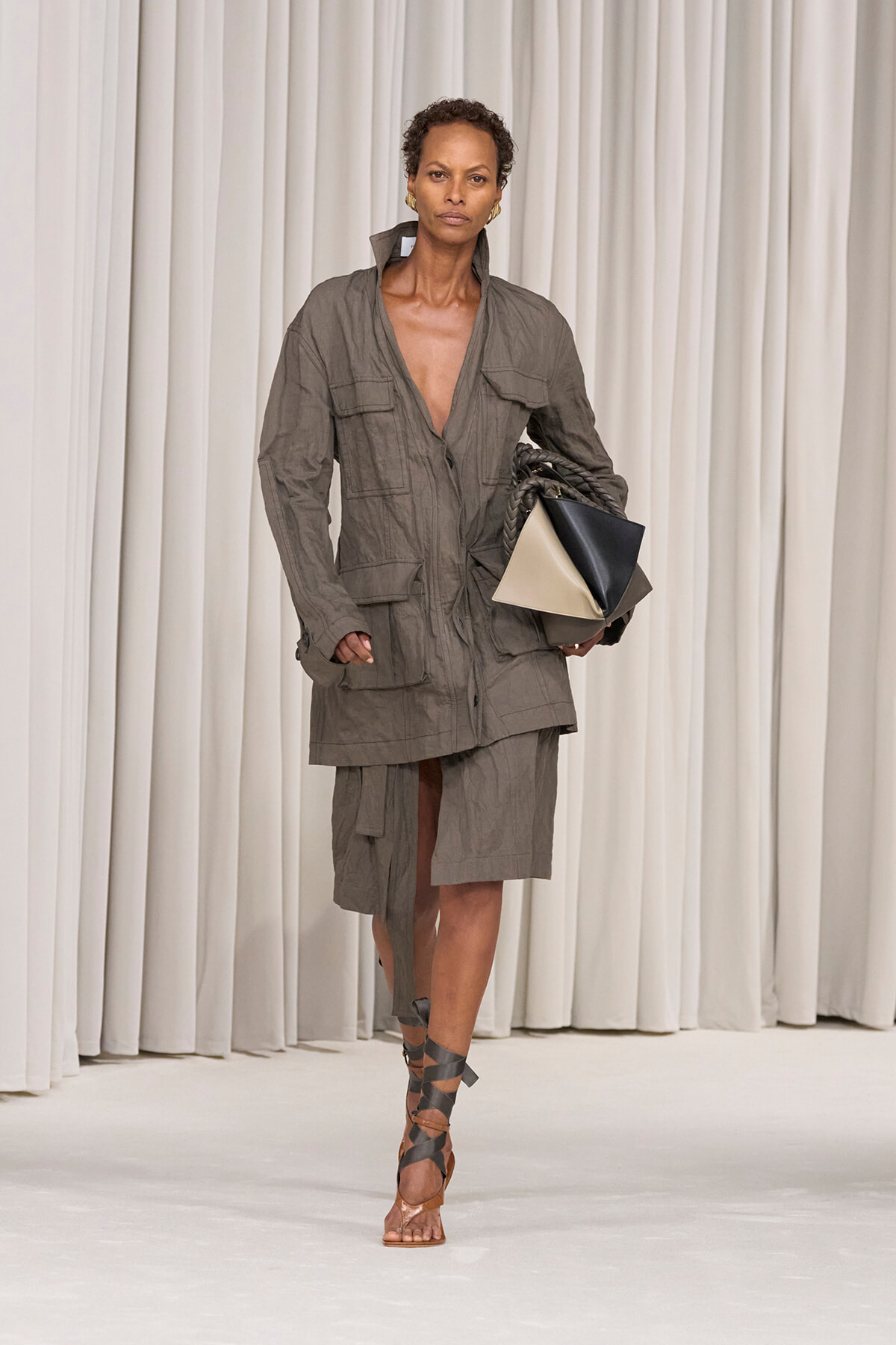 Model walking the runway in a taupe utility jacket and matching skirt, carrying a color-block geometric handbag and wearing strappy sandals.