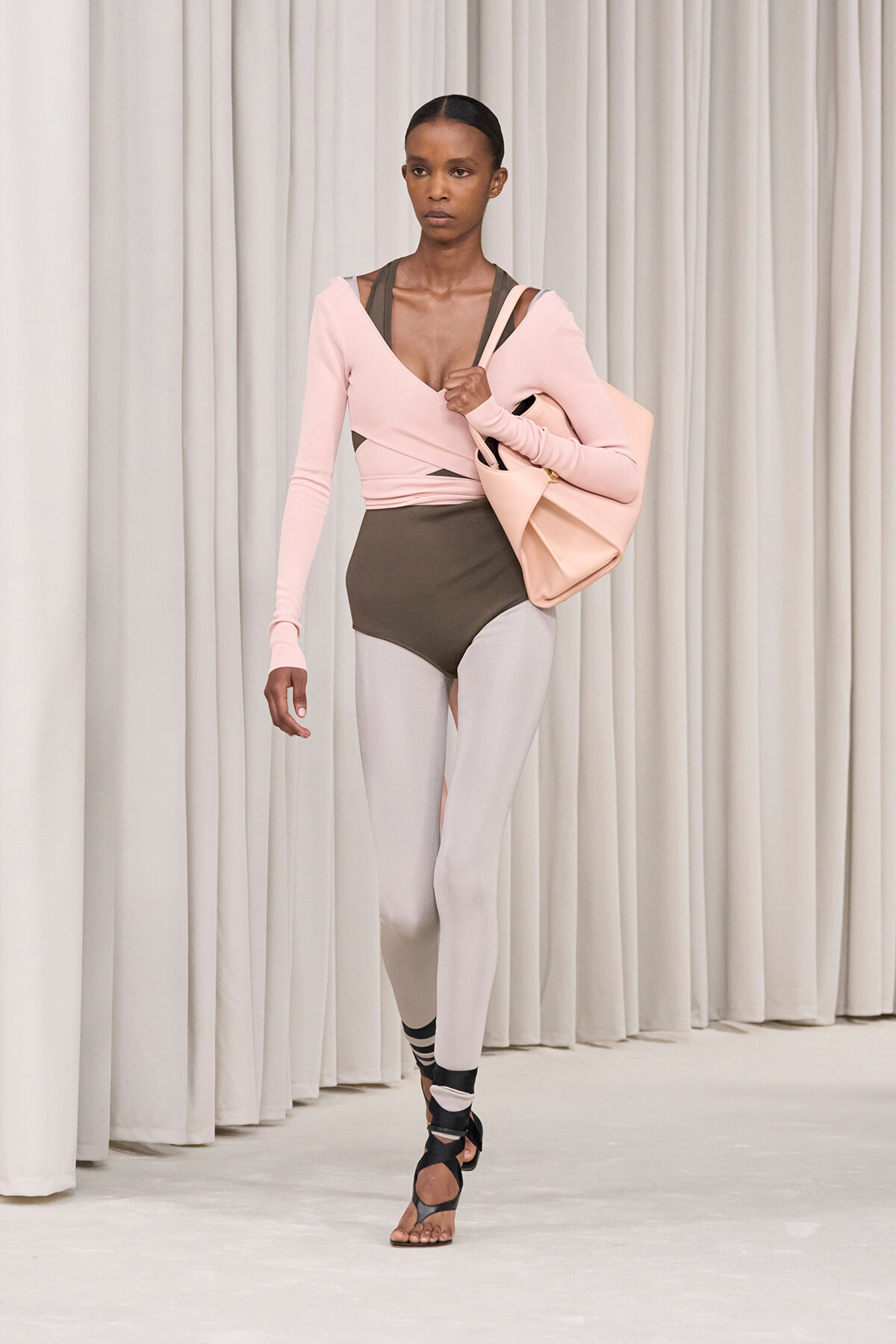 Model walking a runway in a pale pink wrap top over a brown bodysuit, light gray leggings, and black strappy sandals, carrying a large pale pink bag.