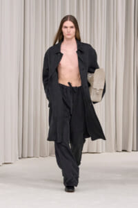 Male runway model in an oversized black coat and matching pants, shirtless, carrying a beige bag against a neutral curtain backdrop.