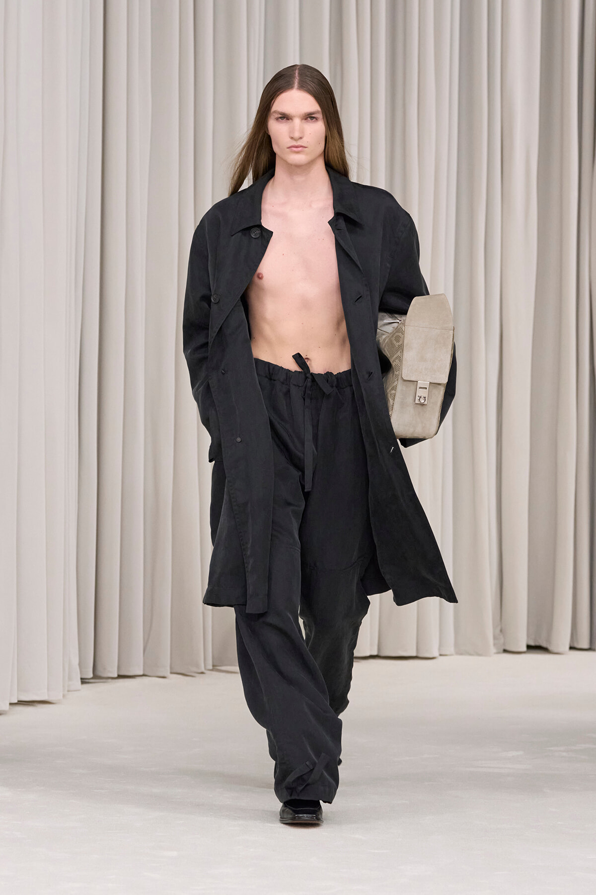 Male runway model in an oversized black coat and matching pants, shirtless, carrying a beige bag against a neutral curtain backdrop.