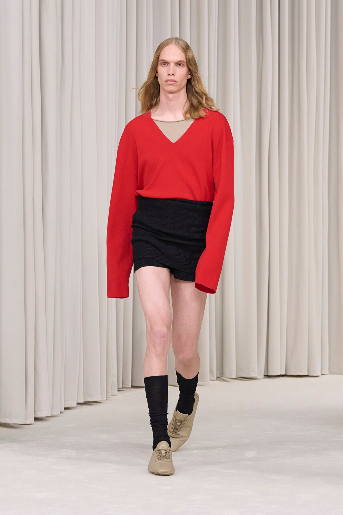 Model walking runway in a red V-neck sweater over a beige top, paired with a black mini skirt, black socks, and tan sneakers against a light curtain backdrop.