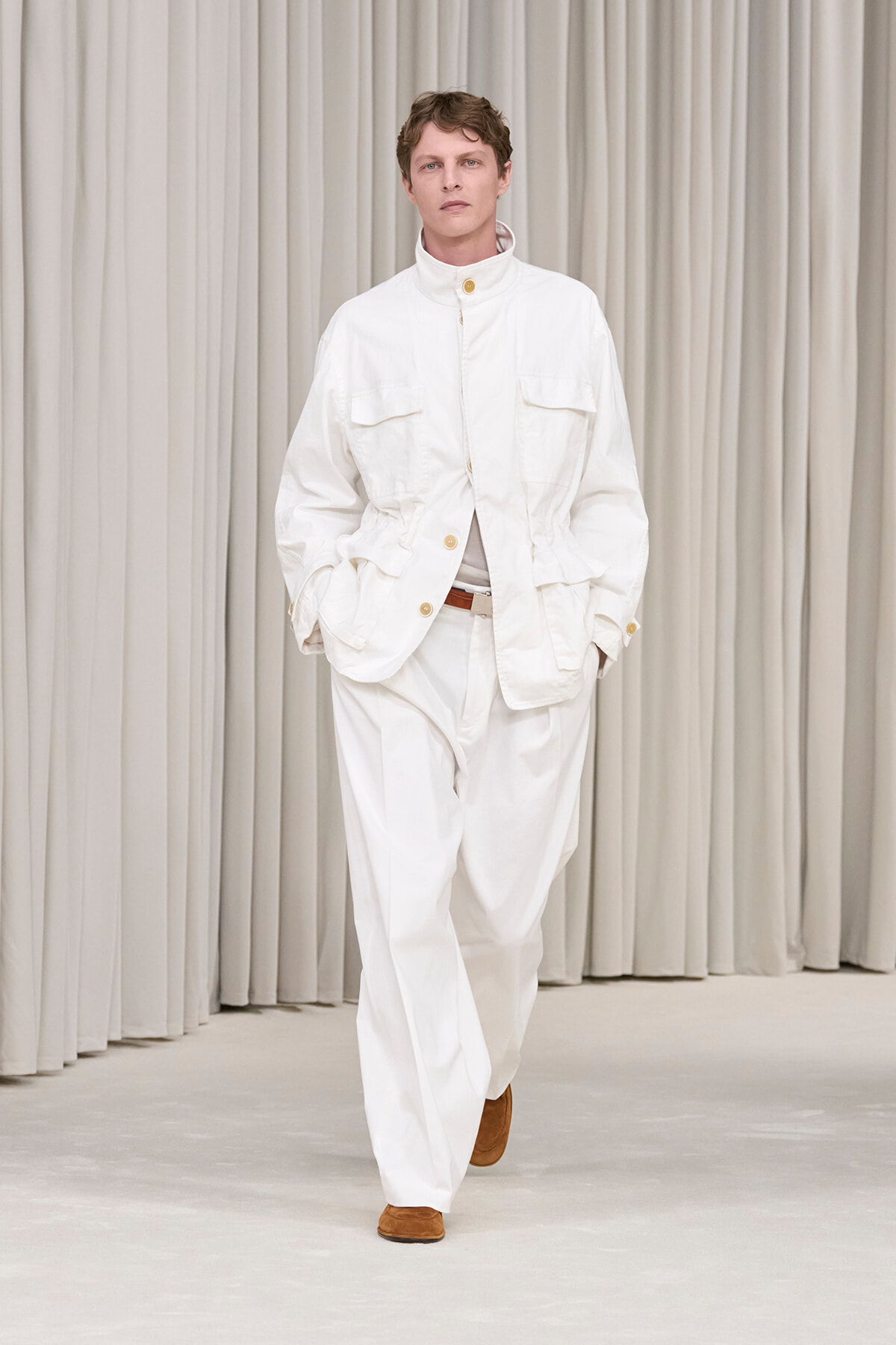 Male model walking a runway in an all-white outfit: high-neck white jacket with large pockets, white pants, and brown shoes against pale curtains.
