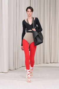 Model walking a runway in a black wrap-style top, taupe bodysuit, red leggings, white wrap sandals, and a large black tote bag.