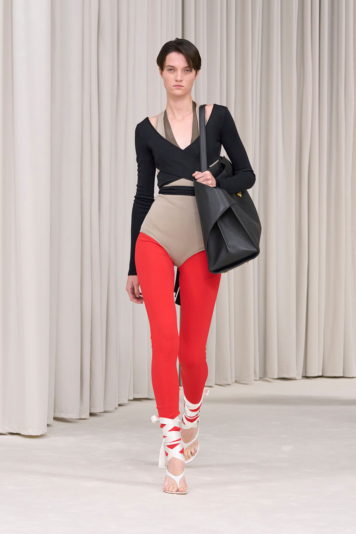 Model walking a runway in a black wrap-style top, taupe bodysuit, red leggings, white wrap sandals, and a large black tote bag.