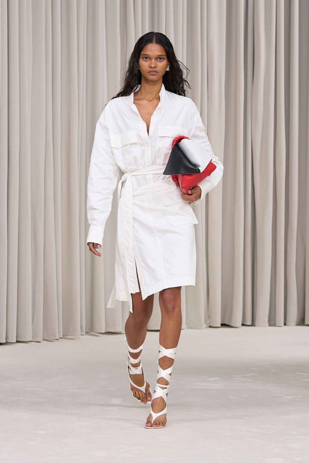 Model walking a white shirt-dress with a belt, holding a red, black, and white clutch on her hip.