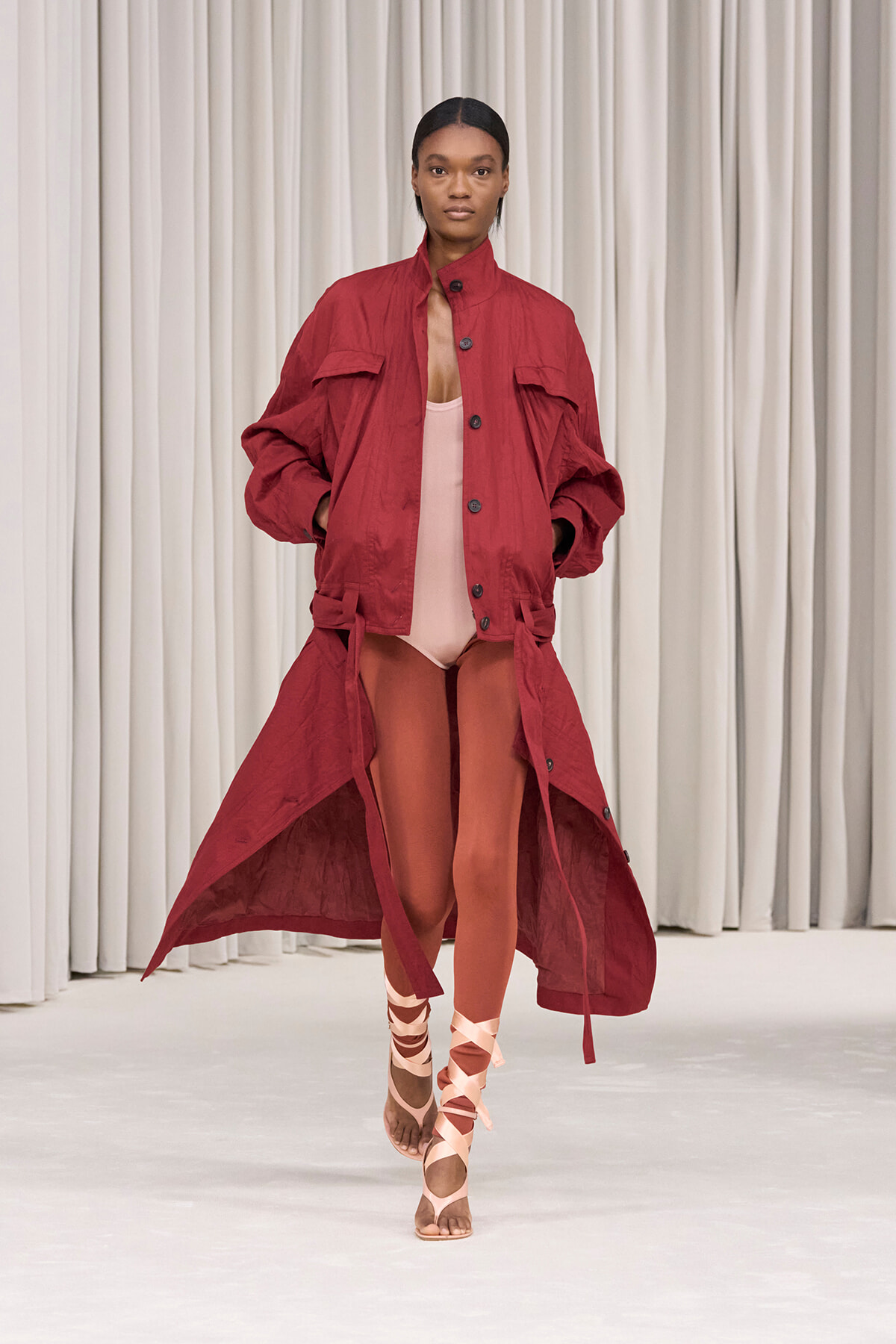 Model walks the runway in a red oversized coat open at the front over a pale pink bodysuit, with crisscross pink sandals.