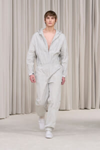Male model wearing a light gray crinkled jumpsuit with a front zipper and hood, walking on a runway.