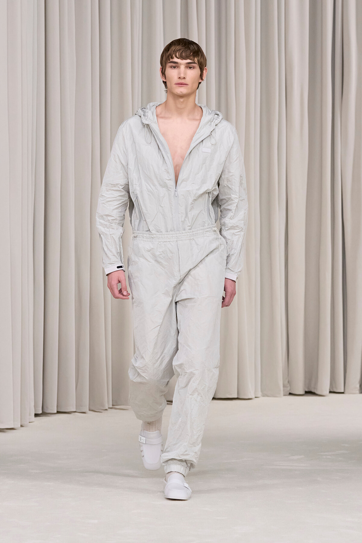 Male model wearing a light gray crinkled jumpsuit with a front zipper and hood, walking on a runway.