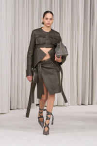 Model in a dark olive leather cropped jacket and asymmetrical wrap skirt with long belt straps, carrying a gray bag, and wearing crisscross ankle-strap heels on a runway.
